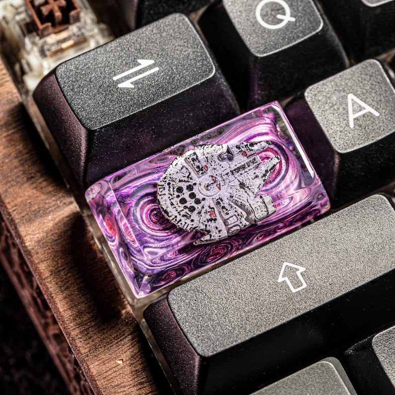 Puede incluir: Un tap&oacute;n de tecla de resina morado y rosa con un dise&ntilde;o de nave espacial Millennium Falcon blanco. El tap&oacute;n de tecla est&aacute; en un teclado negro.