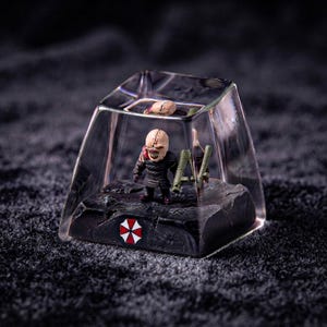 May include: A clear, square resin keycap featuring a miniature Nemesis figure from Resident Evil inside. The figure holds a green rocket launcher, stands on a dark, textured base with a red and white umbrella logo, and has a small brain piece on top.