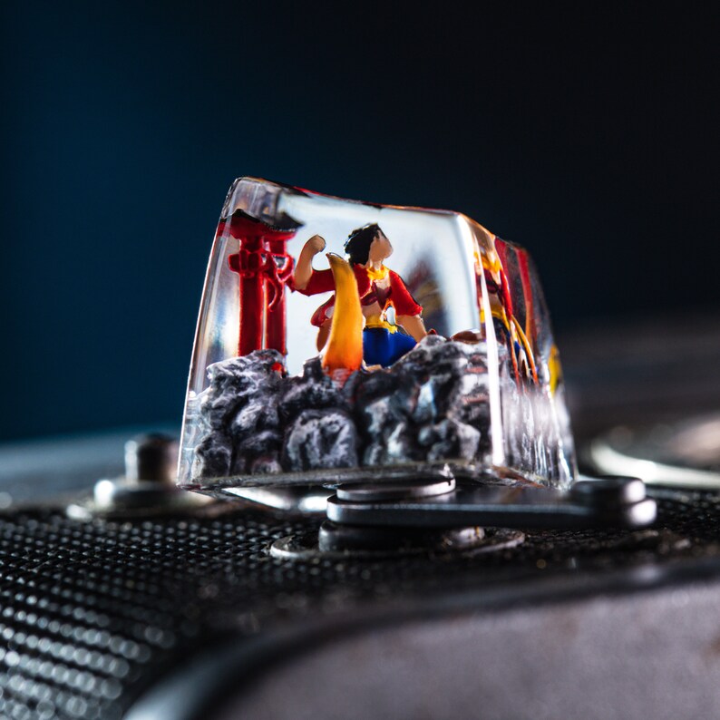 Puede incluir: Un tap&oacute;n de tecla de resina transparente con una figura en miniatura de una persona con un atuendo rojo y amarillo de pie frente a una estructura roja. El tap&oacute;n de tecla est&aacute; en una c&aacute;mara negra.