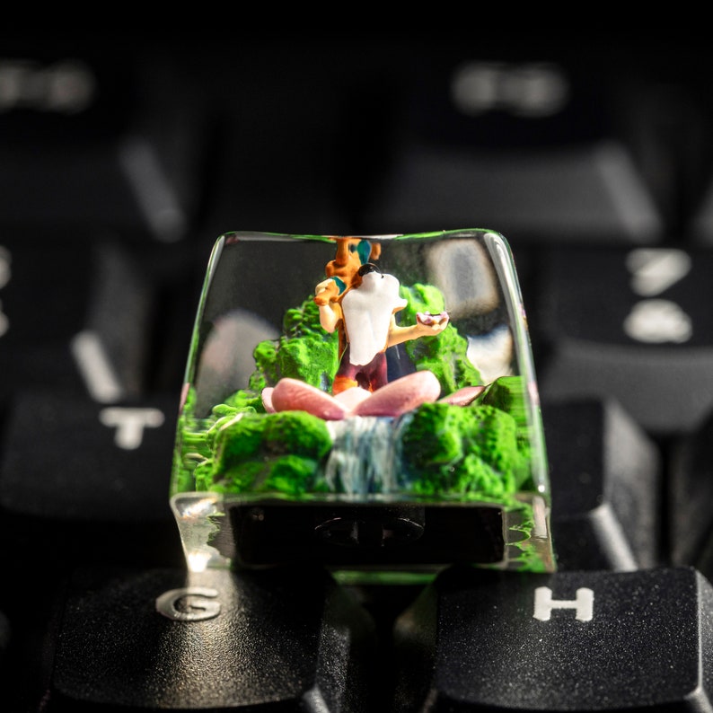 May include: A clear plastic keycap with a miniature figurine of a person sitting on a pink lotus flower in a green and brown landscape. The figurine is surrounded by a waterfall and green foliage.