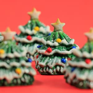 Könnte beinhalten: Kleine Keramik-Weihnachtsbäume in Grün, Weiß und bunten Farben. Jeder Baum ist mit einem Stern gekrönt und mit kleinen, bunten Ornamenten verziert. Die Bäume stehen vor einem roten Hintergrund und schaffen eine festliche Urlaubsszene.