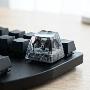 May include: A clear resin mechanical keyboard keycap featuring a miniature dark brown spider-like robot with a single red eye, positioned on a textured gray rocky base, installed among standard black keys on a keyboard.