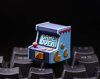 May include: A miniature arcade machine keycap in blue and navy with gold and red accents. The keycap features the words "GAME OVER" on the screen. The keycap is sitting on a black keyboard.