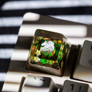 May include: A clear resin keycap with a green and yellow floral design and a white lion logo. The keycap is on a keyboard.