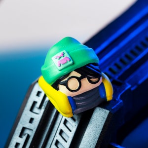 Pode incluir: Uma pequena figura verde, amarela e azul de uma pessoa usando um gorro verde com um logotipo rosa e &oacute;culos de arma&ccedil;&atilde;o preta. A figura est&aacute; usando uma m&aacute;scara preta e tem cabelo preto.