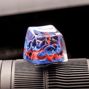 May include: A clear resin keycap with a blue and red abstract design. The design features swirling patterns and a textured surface.