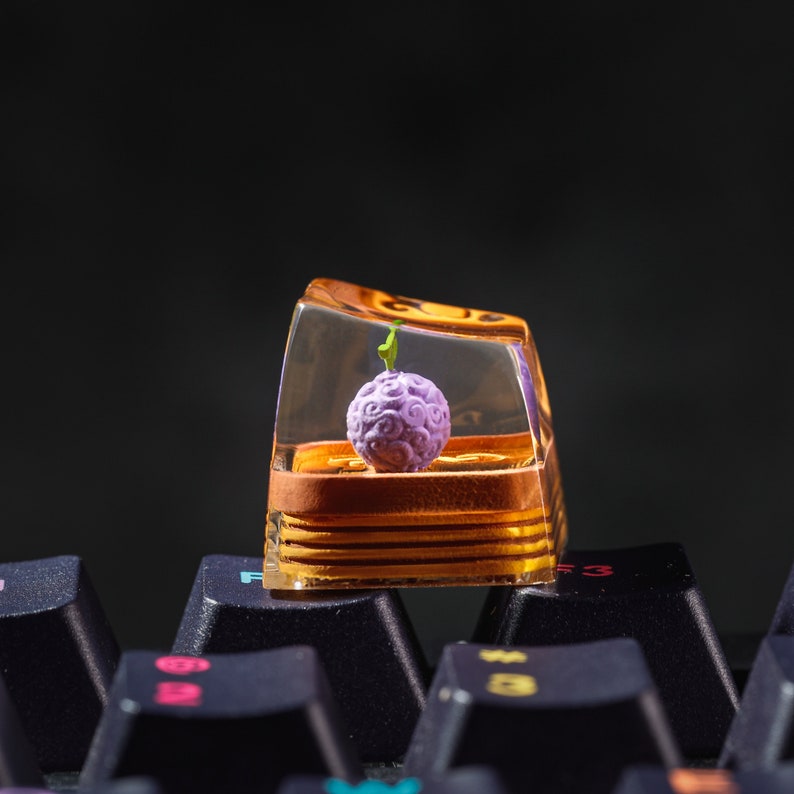 Puede incluir: Un keycap de resina transparente con una fruta morada y verde en su interior. El keycap est&aacute; sobre un teclado negro.