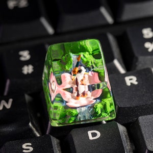 May include: A green, clear, plastic keycap with a miniature figurine of a person inside. The figurine is wearing a white shirt and a pink skirt. The keycap is sitting on a black keyboard.