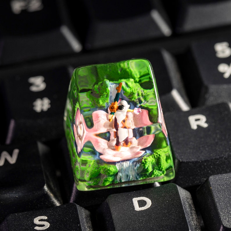 May include: A green, clear, plastic keycap with a miniature figurine of a person inside. The figurine is wearing a white shirt and a pink skirt. The keycap is sitting on a black keyboard.