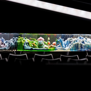 Peut inclure: Un bloc de résine transparente avec une scène de paysage miniature représentant une vague bleue et blanche, une terre verte et brune et des figurines colorées.