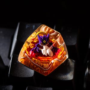 May include: A custom keycap with a purple and white character set within clear, amber resin. The keycap is placed on a black keyboard, showcasing the vivid colours and intricate details of the design.