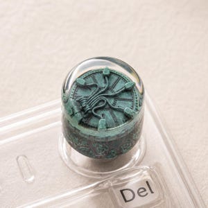 Op de afbeelding: Een ronde mechanische toetsenbordtoets met een groenblauw, verweerd metaalontwerp met een centraal tandwielmotief en stralende lijnen, ingesloten in een heldere, gladde, afgeronde harskoepel, geplaatst boven een doorzichtige toets met Del-tekst.