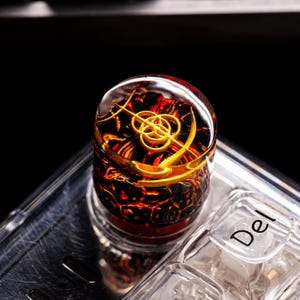 Könnte beinhalten: Eine individuelle Tastenkappe mit einer klaren, abgerundeten Oberseite und einem feurigen, lavaartigen Inneren. Die Tastenkappe hat ein goldfarbenes, stilisiertes Design. Die Tastenkappe befindet sich auf einer Tastatur, wobei die "Entf"-Taste sichtbar ist.