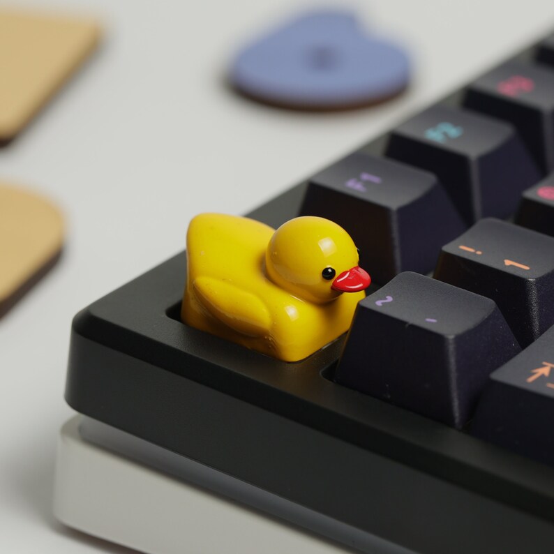 Puede incluir: Un tap&oacute;n de tecla de pato de goma amarillo con un pico rojo y ojos negros se encuentra en un teclado mec&aacute;nico negro.
