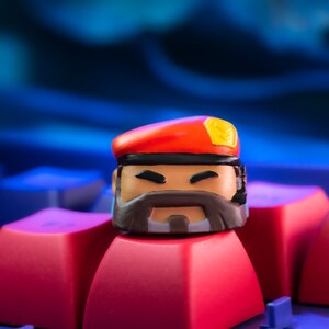 Pode incluir: Um keycap vermelho e amarelo com um rosto de desenho animado, uma barba preta e um bon&eacute; vermelho. O keycap est&aacute; sentado em uma tecla de teclado vermelha.