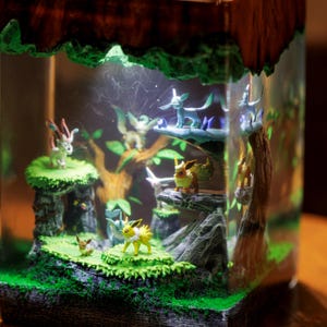 May include: A square resin diorama featuring multiple small Eevee evolution figurines placed on green mossy platforms and rocky terrain. The scene includes miniature trees and internal lighting that illuminates the figures within the clear block.