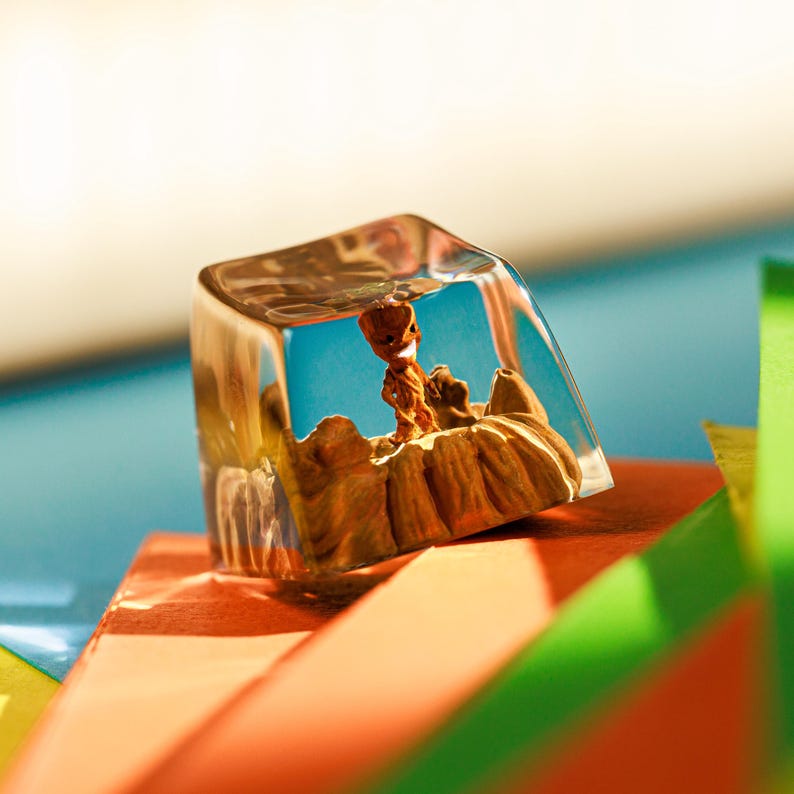 Puede incluir: Un tap&oacute;n de tecla de resina transparente con una figurita en miniatura de una criatura marr&oacute;n con forma de &aacute;rbol en su interior. El tap&oacute;n de tecla est&aacute; sobre una superficie colorida.