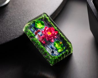 May include: A rectangular, clear resin keycap featuring a miniature pond scene. A vibrant pink lotus flower is surrounded by green lily pads and lush green foliage. The water is a mix of blue and white, creating a tranquil miniature landscape.