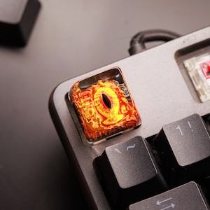 Può includere: Un cappuccio per tasti in resina trasparente con un design a occhio rosso e giallo. Il cappuccio per tasti è su una tastiera meccanica nera.