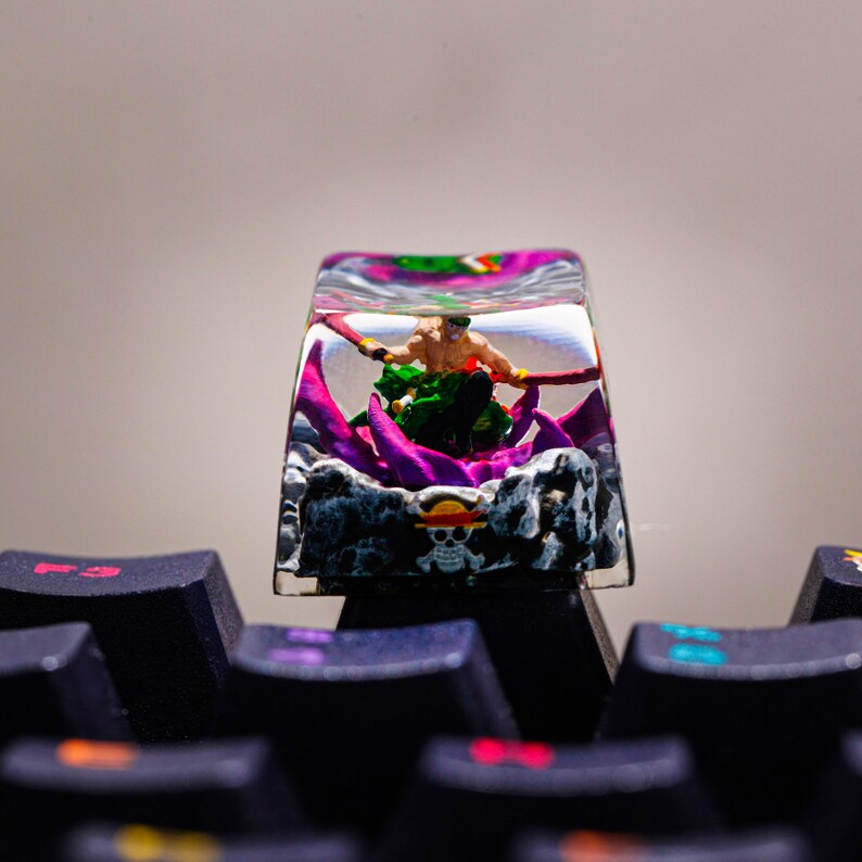 Puede incluir: Un tap&oacute;n de tecla de resina transparente con una figura en miniatura de un personaje del anime One Piece. El personaje sostiene una espada y est&aacute; rodeado de resina morada y verde. El tap&oacute;n de tecla est&aacute; sobre un teclado negro.