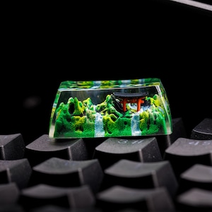 May include: A green and white miniature landscape scene with a red and black torii gate inside a clear resin keycap. The keycap is sitting on a black keyboard.
