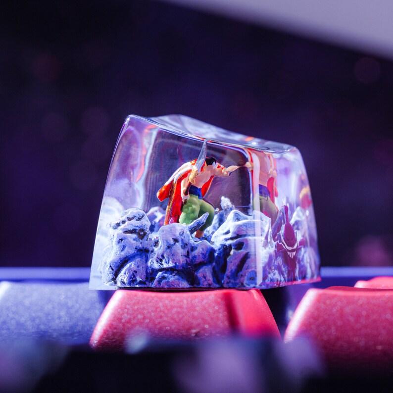 Puede incluir: Un tap&oacute;n de tecla de pl&aacute;stico transparente con una figura en miniatura de una persona con un atuendo rojo y verde en su interior. La figura est&aacute; de pie sobre una superficie rocosa azul y blanca. El tap&oacute;n de tecla est&aacute; sobre un teclado rojo y negro.