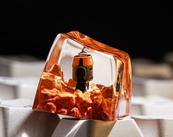May include: A clear resin keycap with a miniature orange and black robot inside. The keycap is sitting on a white keyboard.