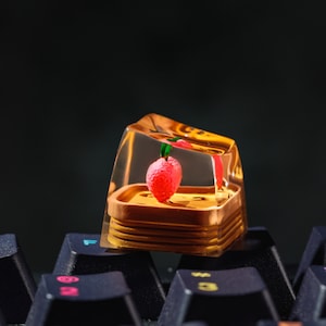 Peut inclure: Un capuchon de touche en résine transparente avec un design en couches présente une petite fraise rose à l'intérieur. Le capuchon de touche est posé sur un clavier noir.