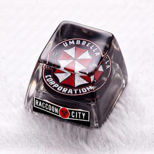 Puede incluir: Una tecla de teclado mecánico negra translúcida con un logotipo circular de Umbrella Corporation con un diseño de paraguas rojo y blanco. El texto Umbrella Corporation rodea el logo y Raccoon City aparece en la parte frontal.