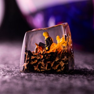 Puede incluir: Un cubo de resina transparente con una figura en miniatura de una persona sentada en un mont&oacute;n de resina amarilla y negra. La resina es texturizada y se asemeja a llamas.