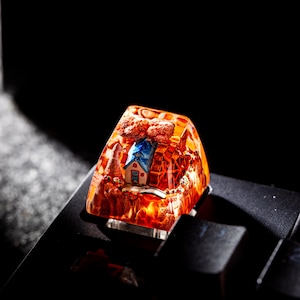 Peut inclure: Une touche de clavier artisanale personnalisée représentant un village miniature. La touche est encapsulée dans une résine transparente de couleur ambre, avec une petite maison, des arbres et un pont. Pour clavier mécanique.