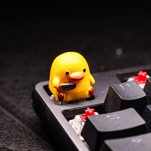 Può includere: Un copritasto a forma di anatra gialla con un coltello nero, seduto su una tastiera nera. Il copritasto ha un design a cartone animato con piedi e becco rossi. I tasti della tastiera sono neri con scritte bianche.
