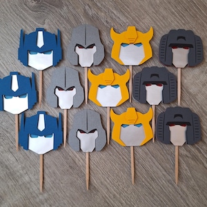 May include: Twelve cupcake toppers featuring Transformers characters. The toppers are made of felt and feature the faces of Optimus Prime, Megatron, Bumblebee, and Starscream in various colors, including blue, yellow, gray, and black.