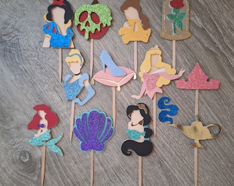 May include: A collection of glittery cupcake toppers featuring various princess-themed designs. Includes Snow White, an apple, Cinderella's slipper, Belle, a rose, Ariel, a seashell, Jasmine, a crown, and a magic lamp. Each topper is mounted on a wooden stick.
