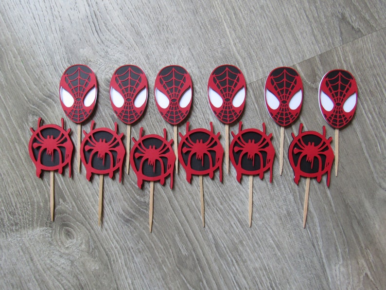 May include: Twelve red and black Spider-Man themed cupcake toppers. Each topper features a red and black Spider-Man mask or a red and black Spider-Man symbol.
