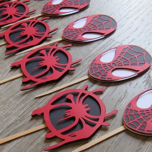 May include: A set of eight red and black Spider-Man themed cupcake toppers. Each topper features a stylised image of Spider-Man's mask with a web design.
