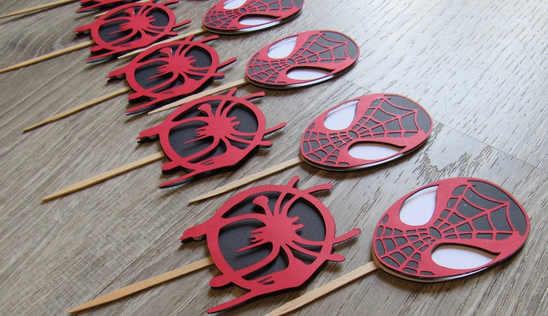 May include: A set of eight red and black Spider-Man themed cupcake toppers. Each topper features a stylised image of Spider-Man's mask with a web design.