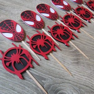 May include: Red and black Spider-Man themed cupcake toppers with a web design. The toppers are on wooden sticks.
