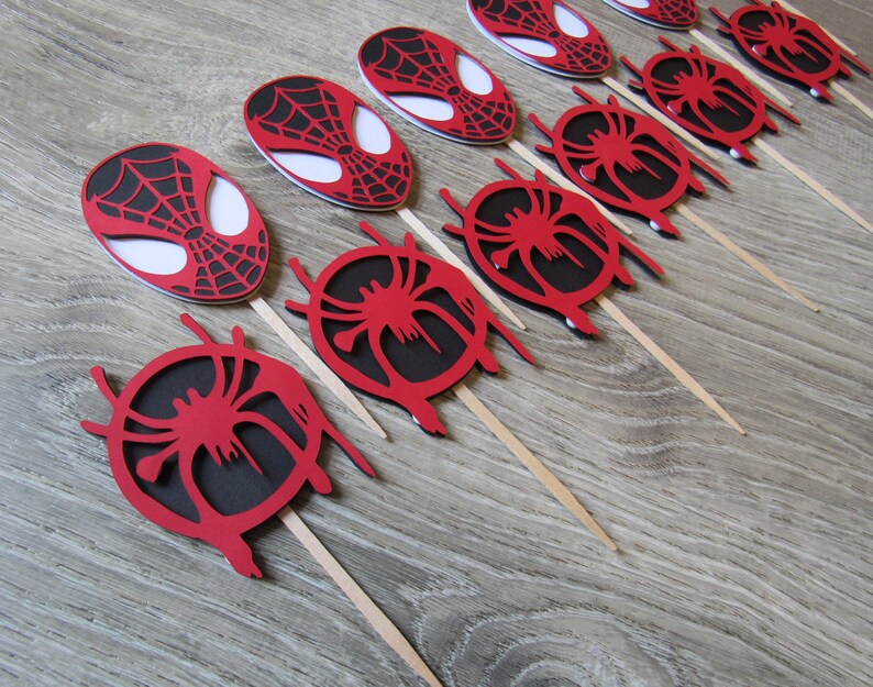May include: Red and black Spider-Man themed cupcake toppers with a web design. The toppers are on wooden sticks.