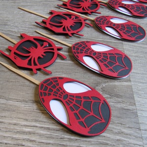 May include: Red and black Spider-Man themed cupcake toppers. Each topper features a red oval with a black web design and a white centre. The toppers are attached to wooden sticks.
