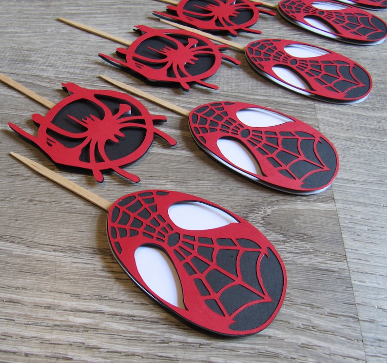 May include: Red and black Spider-Man themed cupcake toppers. Each topper features a red oval with a black web design and a white centre. The toppers are attached to wooden sticks.
