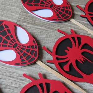 May include: Red and black paper cupcake toppers featuring a Spiderman design. The toppers are on wooden sticks.