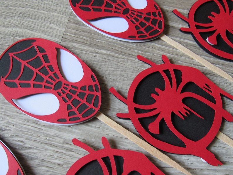 May include: Red and black paper cupcake toppers featuring a Spiderman design. The toppers are on wooden sticks.
