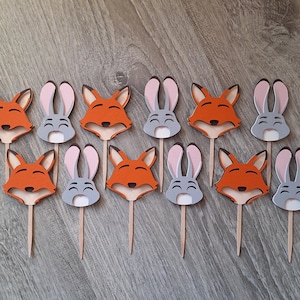 May include: A collection of wooden cupcake toppers featuring fox and bunny designs. The foxes are orange with brown accents, and the bunnies are gray with pink ears. Each topper is attached to a wooden stick.