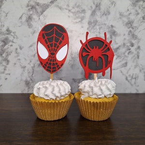 May include: Two cupcakes decorated with red and black Spiderman themed toppers. One topper is a Spiderman mask and the other is a Spiderman web.