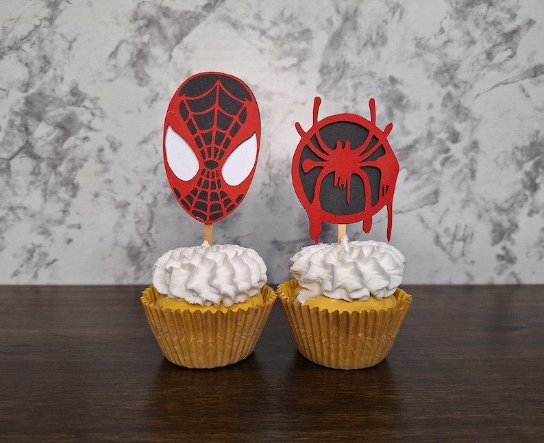 May include: Two cupcakes decorated with red and black Spiderman themed toppers. One topper is a Spiderman mask and the other is a Spiderman web.