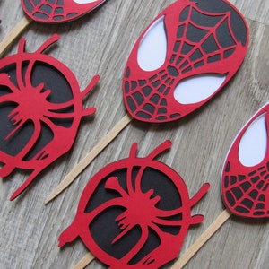 May include: Red and black Spider-Man themed cupcake toppers. The toppers feature a spider web design and a white outline of Spider-Man's face.