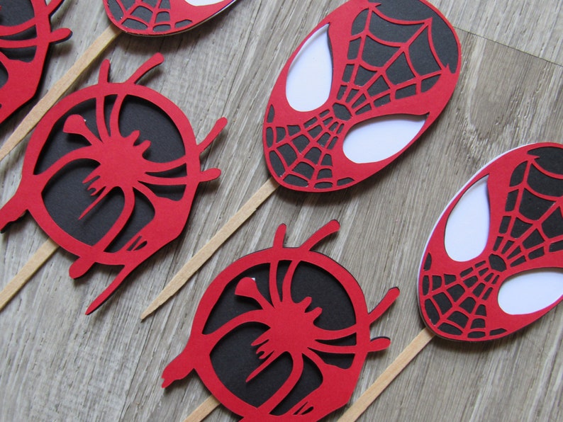 May include: Red and black Spider-Man themed cupcake toppers. The toppers feature a spider web design and a white outline of Spider-Man's face.