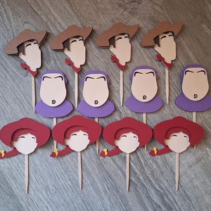 以下が含まれることがあります： 木製の棒に描かれた漫画のキャラクターの紙の切り抜きコレクション。キャラクターには、茶色の帽子をかぶったカウボーイ、紫色の襟のキャラクター、赤い帽子をかぶったカウガールが含まれます。切り抜きはさまざまな色で、パーティーやイベント用である可能性があります。