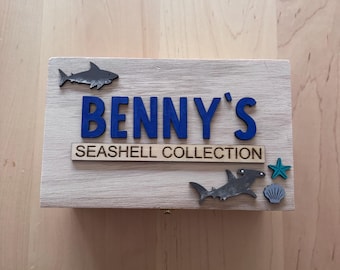 May include: A rectangular light-colored wooden box featuring the blue text BENNY'S and the black text SEASHELL COLLECTION. The lid is decorated with a dark gray shark, a dark gray hammerhead shark, a teal starfish, and a small gray seashell.